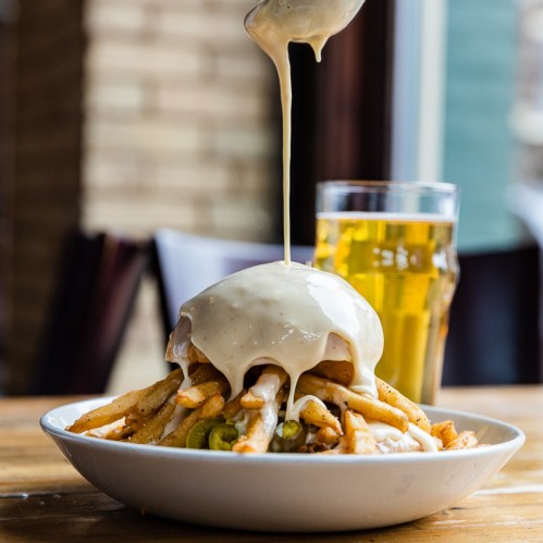 A plate of fries topped with gravy and cheese sauce, with a glass of beer in the background.