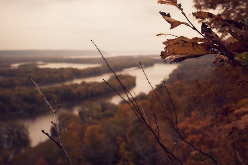 A scenic view of a winding river surrounded by autumn foliage and distant hills under a cloudy sky.