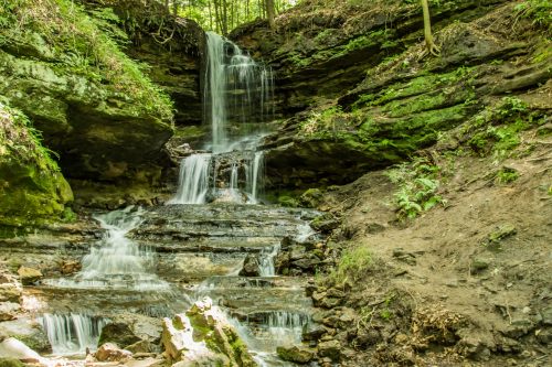 A serene waterfall cascading over rocky layers, surrounded by lush greenery and forested terrain.