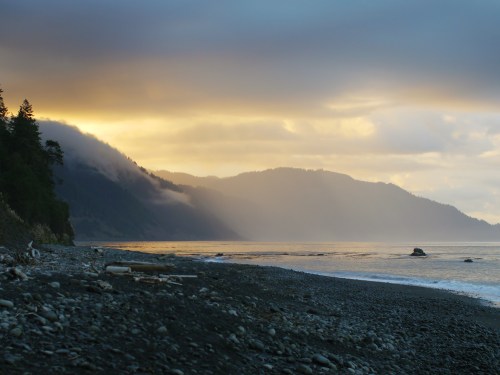 A serene beach scene at sunset, with misty mountains in the background and gentle waves lapping at the shore.