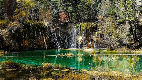 A serene landscape featuring a turquoise lake, a waterfall, and lush greenery surrounded by autumn foliage.
