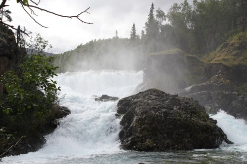 A cascading waterfall surrounded by lush greenery and mist, with large rocks in the foreground.