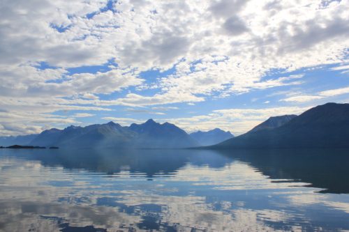 Serene landscape of mountains reflected in calm water under a cloudy blue sky.