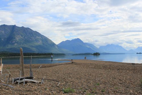 A serene lakeside scene with mountains in the background, featuring a rocky shore and two people in the distance.
