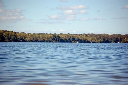 A calm lake reflects the sky, with trees lining the shore and a distant tower visible among the greenery.