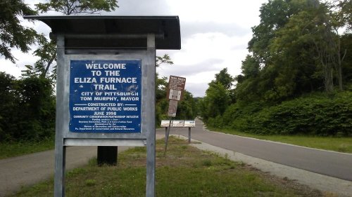 Sign welcoming visitors to the Eliza Furnace Trail in Pittsburgh, with a paved path and greenery in the background.