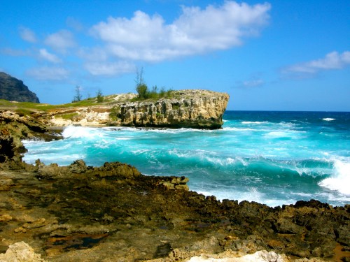 A rocky coastline with turquoise waves crashing against cliffs under a bright blue sky with scattered clouds.