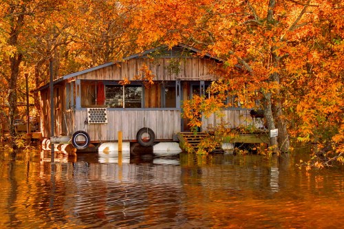 A rustic wooden house surrounded by vibrant autumn foliage, reflecting in calm water.