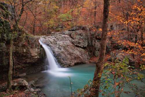 A serene waterfall cascades into a turquoise pool, surrounded by autumn foliage and rocky terrain.