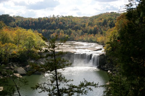A serene waterfall cascades into a calm lake, surrounded by vibrant autumn foliage and lush green trees.