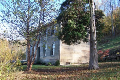 A stone building partially obscured by trees, set in a grassy area with autumn foliage in the background.