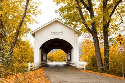 A white covered bridge surrounded by vibrant autumn foliage and a warning sign for weight limit.