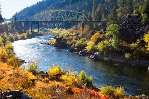 A scenic river flows under a yellow bridge, surrounded by colorful autumn foliage and rocky terrain.