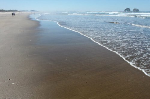 A sandy beach with gentle waves lapping at the shore, people walking in the distance under a clear blue sky.