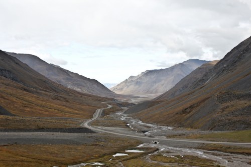 A winding road through a vast, mountainous valley with rocky terrain and patches of grass under a cloudy sky.