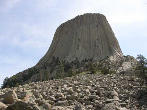 A towering rock formation rises above a rocky landscape, surrounded by sparse trees under a clear blue sky.