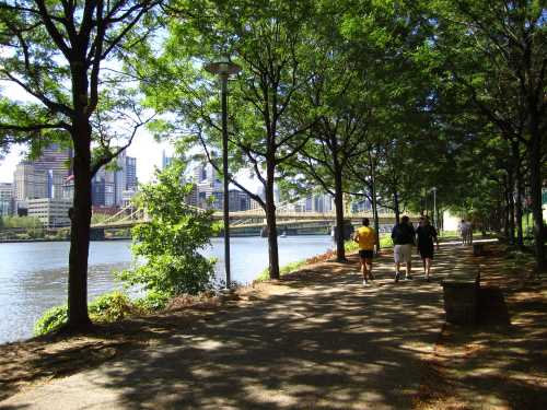 A scenic riverside path lined with trees, people walking, and a bridge visible in the background against a city skyline.