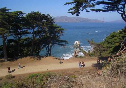 A scenic view of a coastal path with people walking, surrounded by trees and overlooking the ocean and distant hills.