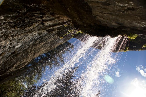 A waterfall cascades down rocky cliffs, surrounded by trees and a bright blue sky with scattered clouds.