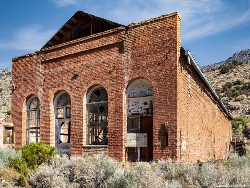 Abandoned brick building with arched windows, surrounded by dry vegetation and mountains in the background.