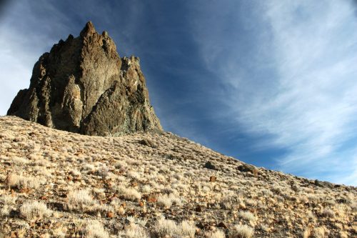 A rugged rock formation rises from a dry, grassy landscape under a blue sky with wispy clouds.