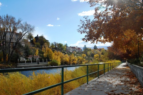 A scenic riverside path lined with autumn trees and colorful foliage under a bright blue sky.