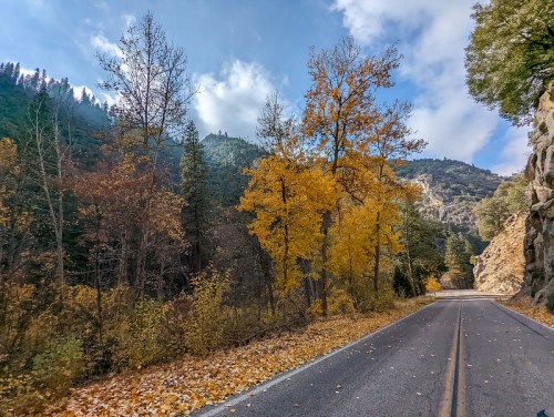 A scenic road lined with autumn trees, surrounded by mountains under a partly cloudy sky.