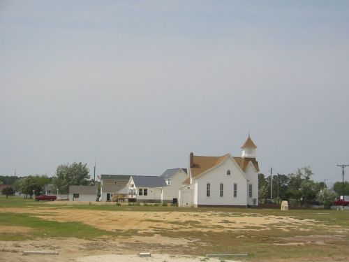 A white church with a steeple stands beside a grassy field, surrounded by a few houses and trees under a cloudy sky.
