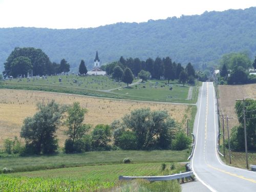A winding road leads to a church surrounded by fields and hills, with a cemetery visible in the background.