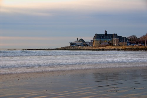 A calm beach scene at dusk, featuring a stone building near the shore and gentle waves lapping at the sand.