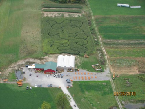 Aerial view of a farm with a corn maze, storage buildings, and parked vehicles surrounded by green fields.