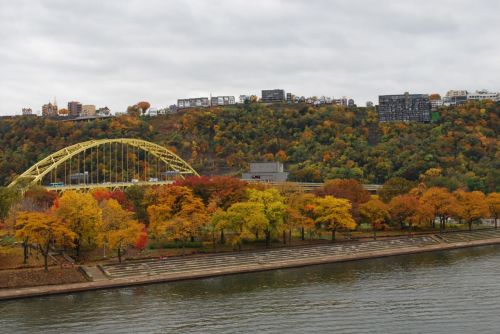 A view of a riverbank with colorful autumn trees and a yellow bridge against a hilly backdrop of buildings.