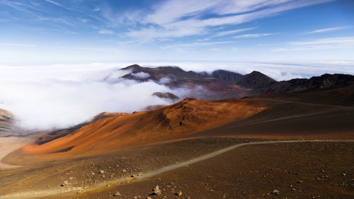 A scenic view of mountains with orange and brown terrain, partially covered by clouds under a blue sky.