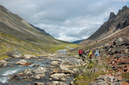Hikers trek along a rocky riverbank in a scenic valley surrounded by mountains under a cloudy sky.
