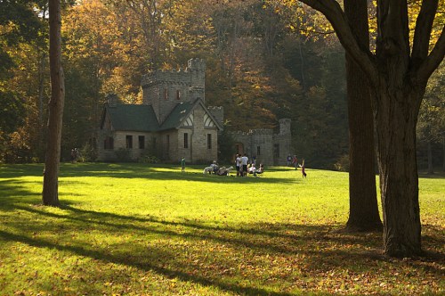 A stone castle surrounded by trees and a grassy area, with people gathered outside in a sunny autumn setting.