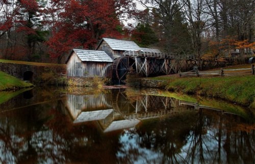 A serene landscape featuring a wooden mill and bridge reflected in calm water, surrounded by autumn foliage.
