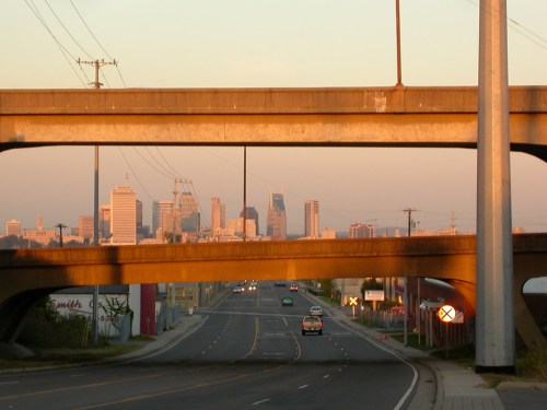 View of a city skyline framed by overpasses at sunset, with a road and cars in the foreground.