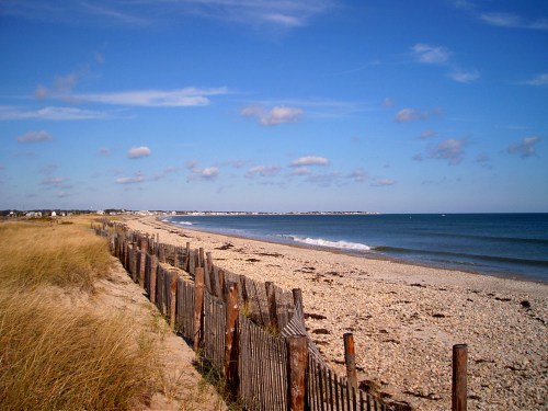 A serene beach scene with a wooden fence, sandy dunes, and gentle waves under a blue sky with scattered clouds.