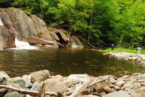 A serene river scene with a small waterfall, surrounded by lush greenery and rocky shores. Two people are near the water.