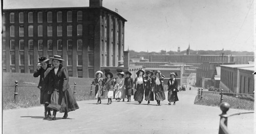 A group of women in early 20th-century attire walking down a road, with industrial buildings in the background.
