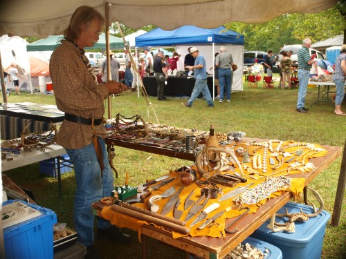A vendor at a market displays handmade jewelry and crafts on a table under a tent, with people browsing in the background.
