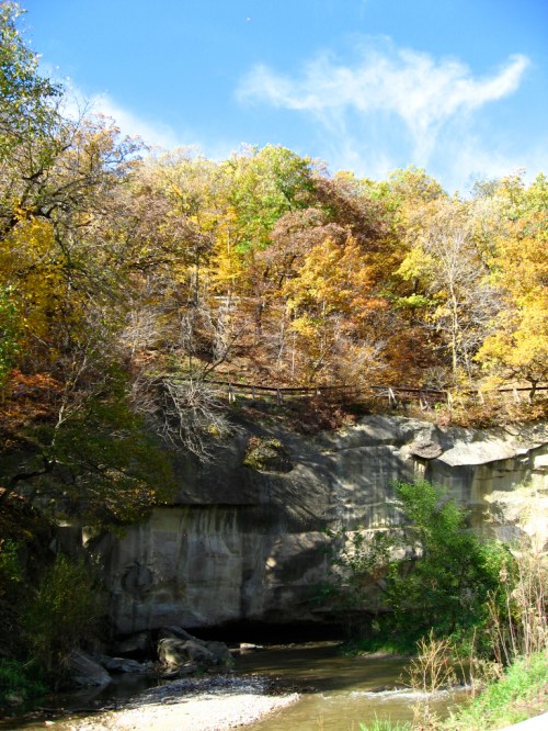 A serene landscape featuring a rocky cliff beside a calm stream, surrounded by vibrant autumn foliage and a clear blue sky.