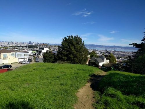 A grassy hilltop view overlooking a cityscape with buildings and a bay under a clear blue sky.