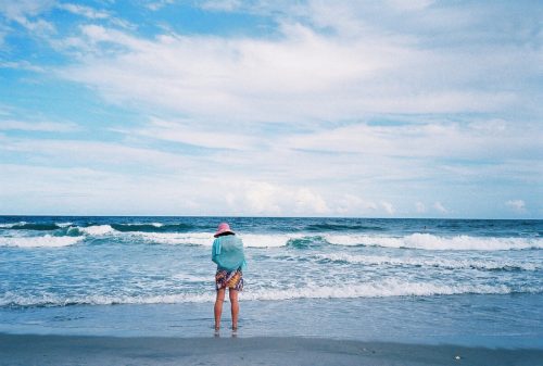 A person in a pink hat stands at the beach, gazing at the ocean under a blue sky with scattered clouds.