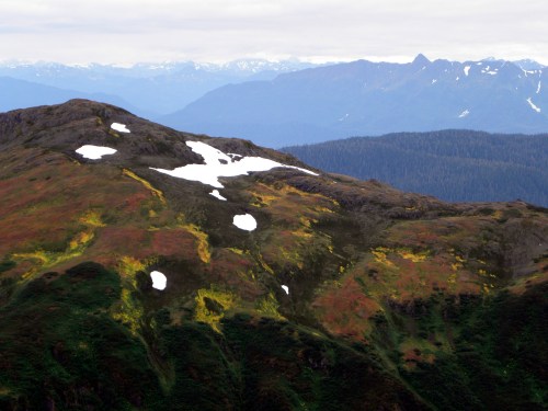 A mountainous landscape with patches of snow and colorful vegetation under a cloudy sky.