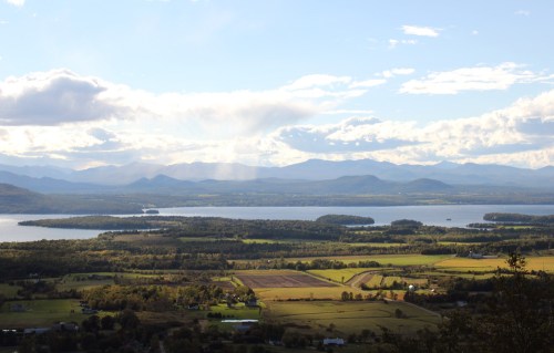 A scenic view of a lake surrounded by green fields and mountains under a partly cloudy sky.
