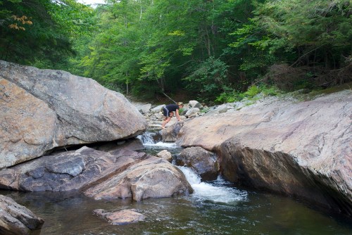 A person crouches by a rocky stream in a lush green forest, surrounded by large boulders and flowing water.