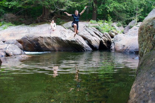 A child jumps into a clear pool of water surrounded by rocks, while another child sits nearby on a rock.