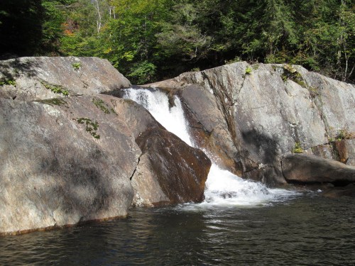 A small waterfall cascading over large rocks into a calm pool, surrounded by lush green trees.