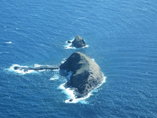 Aerial view of two rocky islands surrounded by deep blue ocean waters. Waves gently crash against the shorelines.